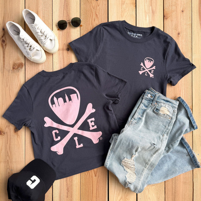 A styled flat-lay of a navy graphic T-shirt set on a light wood plank background, accompanied by light-wash distressed denim shorts, white sneakers, sunglasses, and a black cap with a white logo.