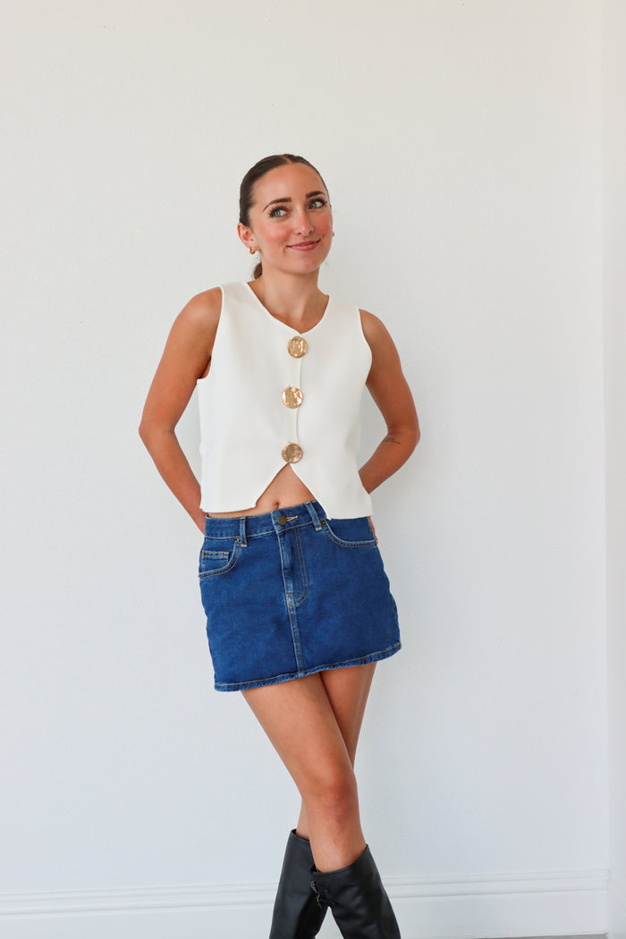 Woman wearing a white sleeveless top with gold buttons, blue denim skirt, and black knee-high boots against a white background.