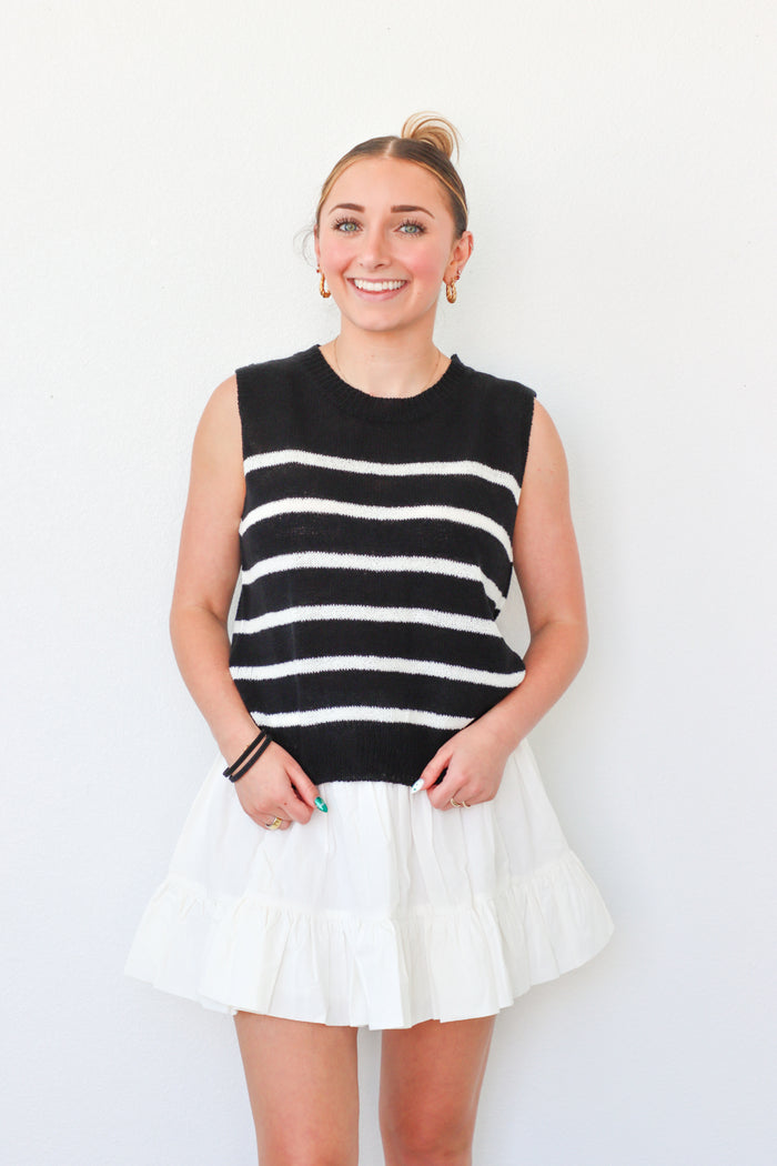 Woman wearing a black and white striped sleeveless sweater with a white skirt on a plain background