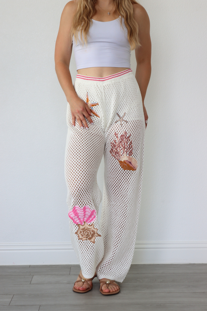 Woman wearing a white crochet pants with shell designs against a plain background