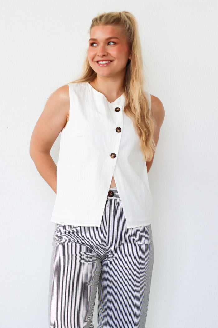 Woman wearing a white sleeveless button-up top with black buttons and striped pants on a white background