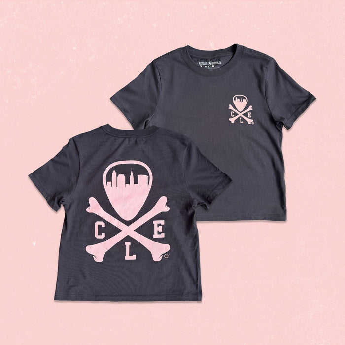 Two dark charcoal gray short-sleeve t-shirts displayed flat against a light pink textured background. The front shirt shows a small light pink skull and crossbones graphic on the left chest, while the back shirt features a large light pink graphic of a shield with a city skyline, crossed bones, and the letters 'C L E'.