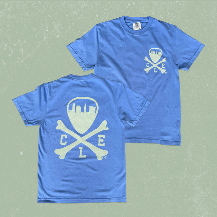 A blue short-sleeve t-shirt shown from both front and back on a textured sage green background. The front features a small white chest graphic of a shield with a city skyline and crossed bones with the letters 'C L E'.