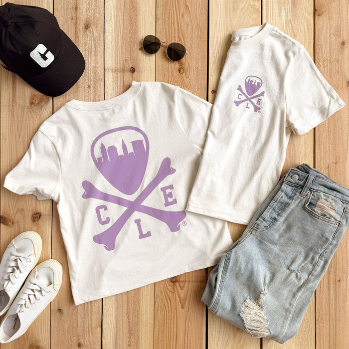A styled flat-lay of a white short-sleeve T-shirt set on a light wood plank background. The set includes a larger back view of the shirt with a purple shield, crossbones, and 'C L E' graphic, and a smaller front view showing a matching purple chest graphic.