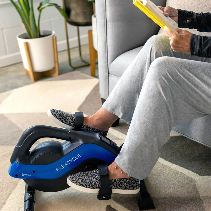 Person reading a book on the couch while pedaling the FlexCycle Plus under-desk bike with feet strapped into the pedals.