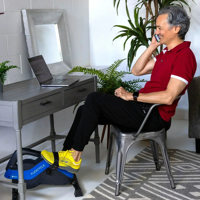 Man on the phone at his desk pedaling the FlexCycle Plus under-desk bike with yellow sneakers while he works.