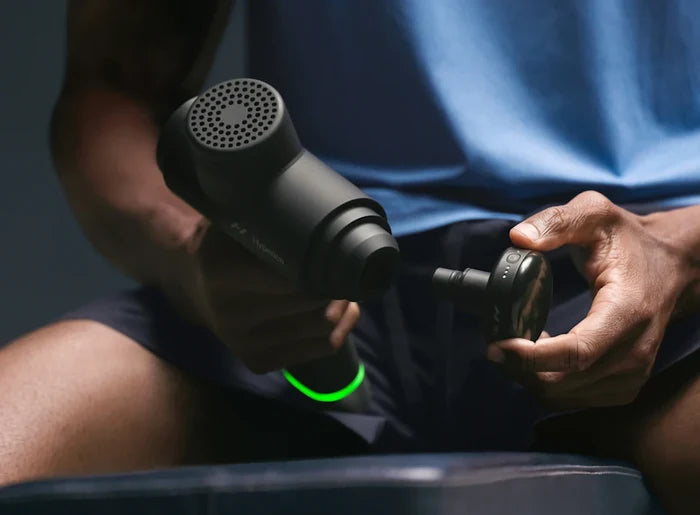 A person holding a black percussion massage device with a detachable Heated Head Attachment | Hyperice, preparing for a muscle recovery session focused on wellness and body care.