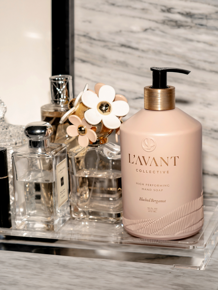 High Performing Hand Soap - Blushed Bergamot | L'AVANT Collective in a blush pink pump bottle displayed on a marble countertop alongside elegant perfume bottles, creating a spa-like skincare setting.