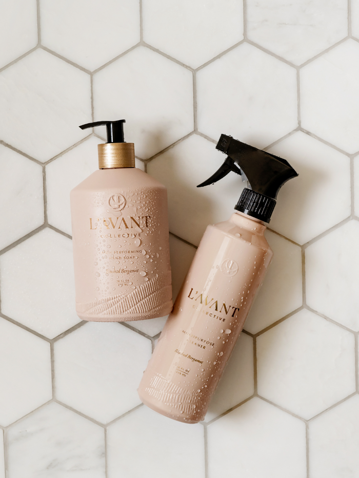 Two blush-colored skincare bottles with water droplets, featuring the High Performing Hand Soap - Blushed Bergamot | L'AVANT Collective, displayed on a white hexagonal tile surface.