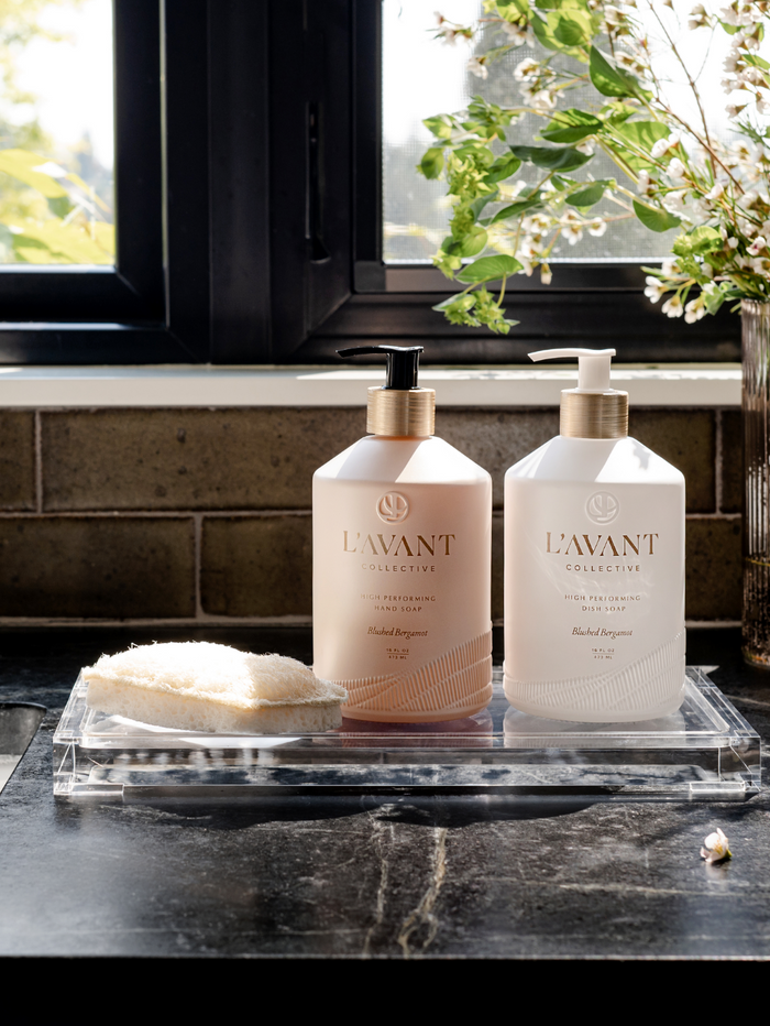 High Performing Hand Soap - Blushed Bergamot | L'AVANT Collective bottle with pump dispenser on a clear tray next to a natural sponge by a sunlit window with greenery in the background.