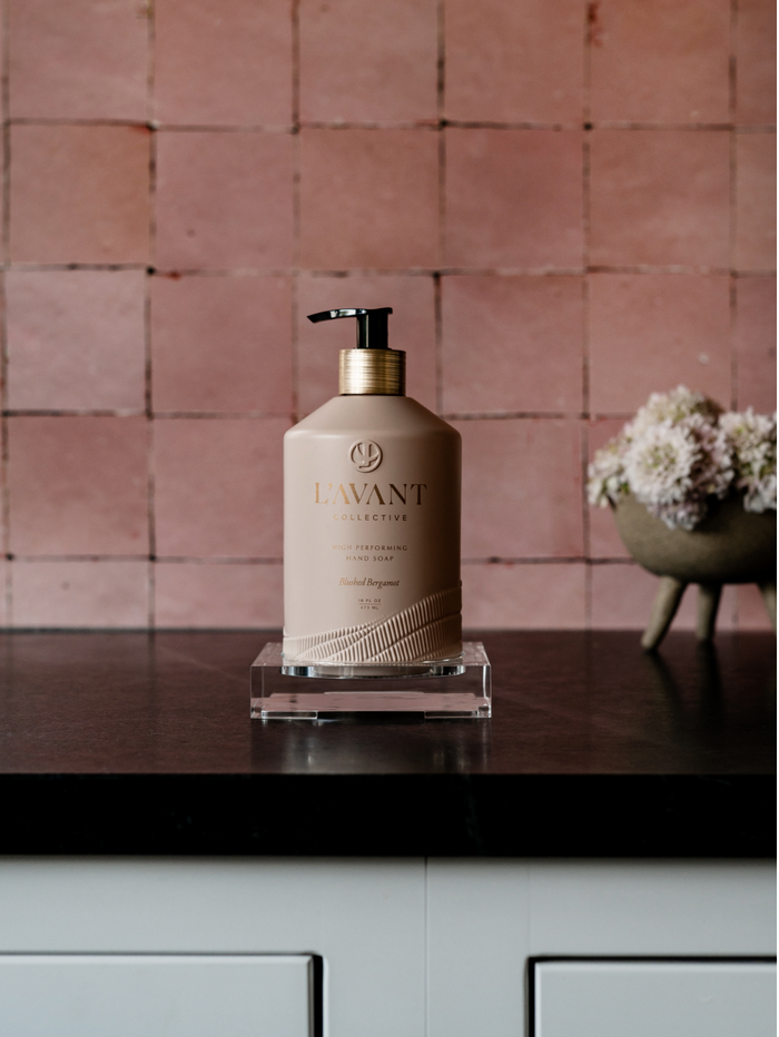 High Performing Hand Soap - Blushed Bergamot | L'AVANT Collective in beige pump bottle on dark countertop with pink tiled wall and floral decor, promoting skincare and wellness.