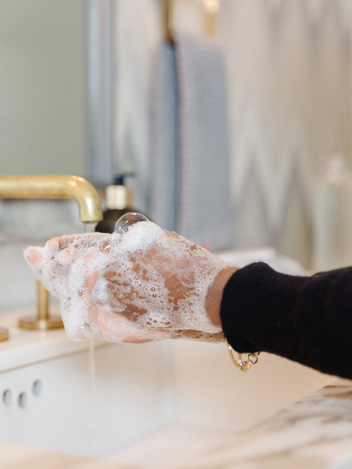 Hands covered in foamy soap lather being washed under running water at a modern bathroom sink, demonstrating use of High Performing Hand Soap - Fresh Linen | L'AVANT Collective for skincare and body care.