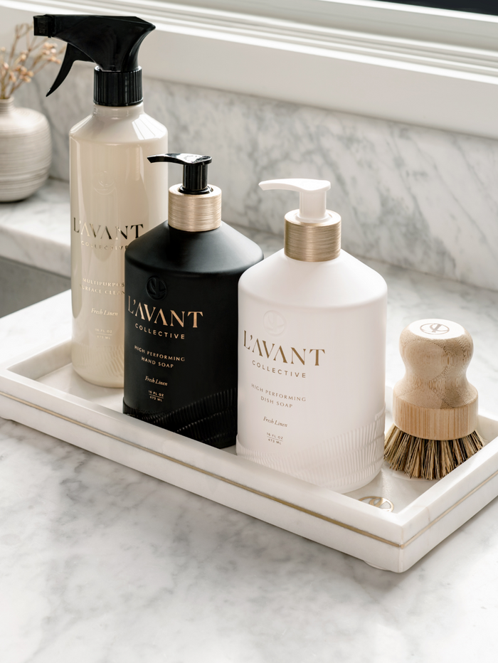 A marble tray holding a black bottle of High Performing Hand Soap - Fresh Linen | L'AVANT Collective, a white bottle of dish soap, and a wooden scrubbing brush, displayed on a marble countertop.