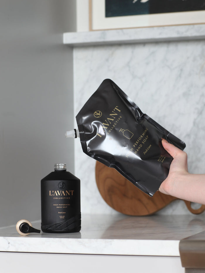 Refilling a sleek black bottle of High Performing Hand Soap - Fresh Linen | L'AVANT Collective on a marble countertop in a modern kitchen setting, highlighting premium skincare and body care.