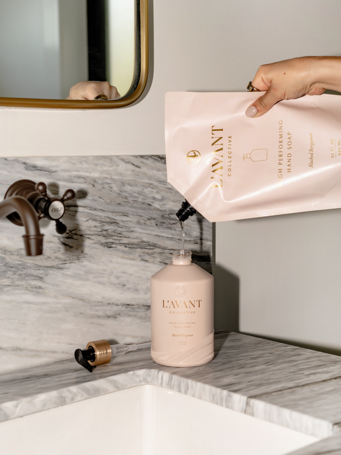 Hand pouring liquid soap from a pink refill pouch into a pink bottle on a marble countertop, featuring High Performing Hand Soap Refills | L'AVANT Collective.