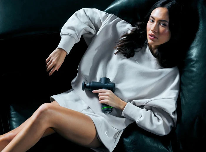 Woman reclining on a dark leather couch using the Hypervolt Go 2 | Hyperice percussion massage device for muscle recovery and wellness.