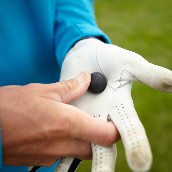 Close-up of a hand wearing a white glove pressing a small black wellness device called Micro Rounds | RAD on the back of the glove, outdoors with green grass in the background.