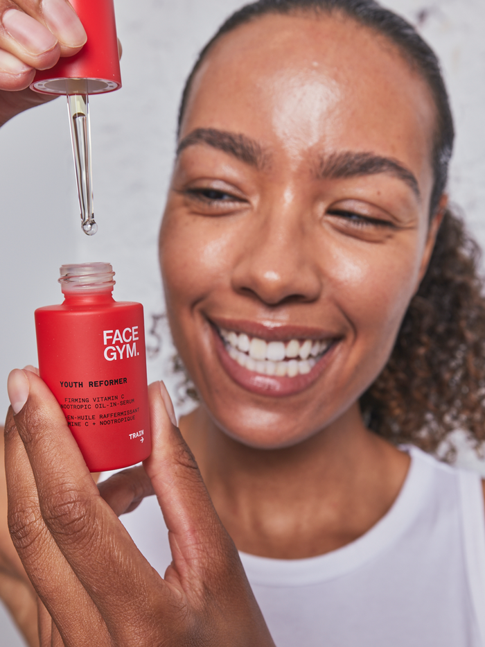 a woman smiling and holding a dropper over a small red bottle