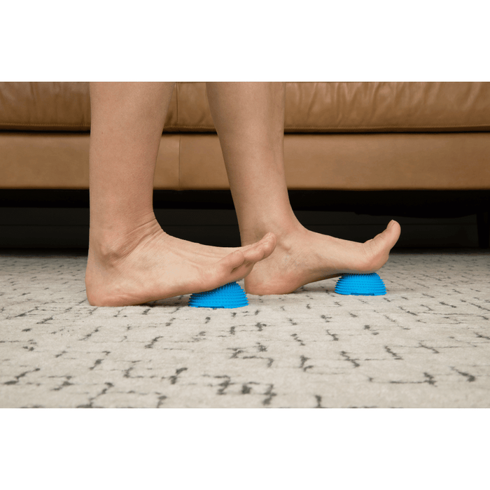 Person using blue textured Neuro Ball | RAD devices under bare feet on a carpeted floor, demonstrating a foot massage and recovery tool for wellness and body care.
