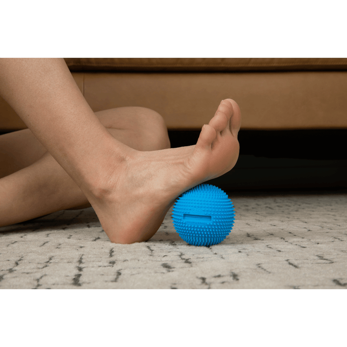 Person using a blue textured Neuro Ball | RAD under the foot for targeted muscle relief and recovery on a carpeted floor near a brown couch.