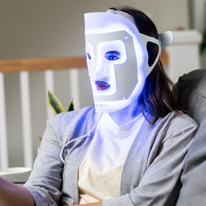 Woman sitting outdoors wearing the red light therapy face mask lit up blue, with its cord running to a handheld controller.