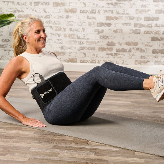 Older woman doing a seated leg-raise ab exercise on a yoga mat while wearing the red light therapy waist wrap.