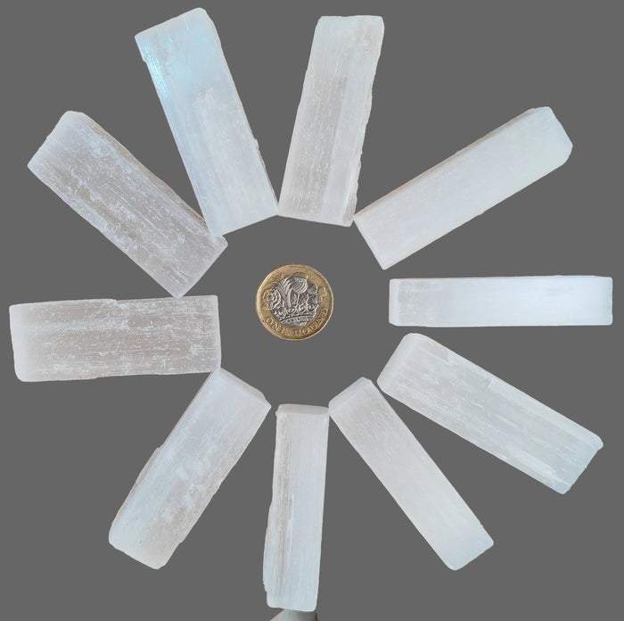 Nine translucent white selenite crystal sticks arranged in a circular pattern on a wooden surface, with a British coin placed in the center for scale.