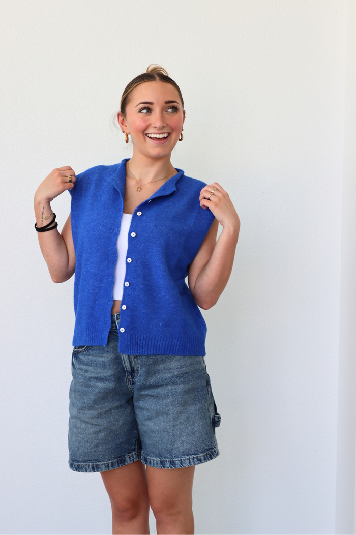Woman wearing a blue knitted vest over a white shirt and denim shorts on a plain background