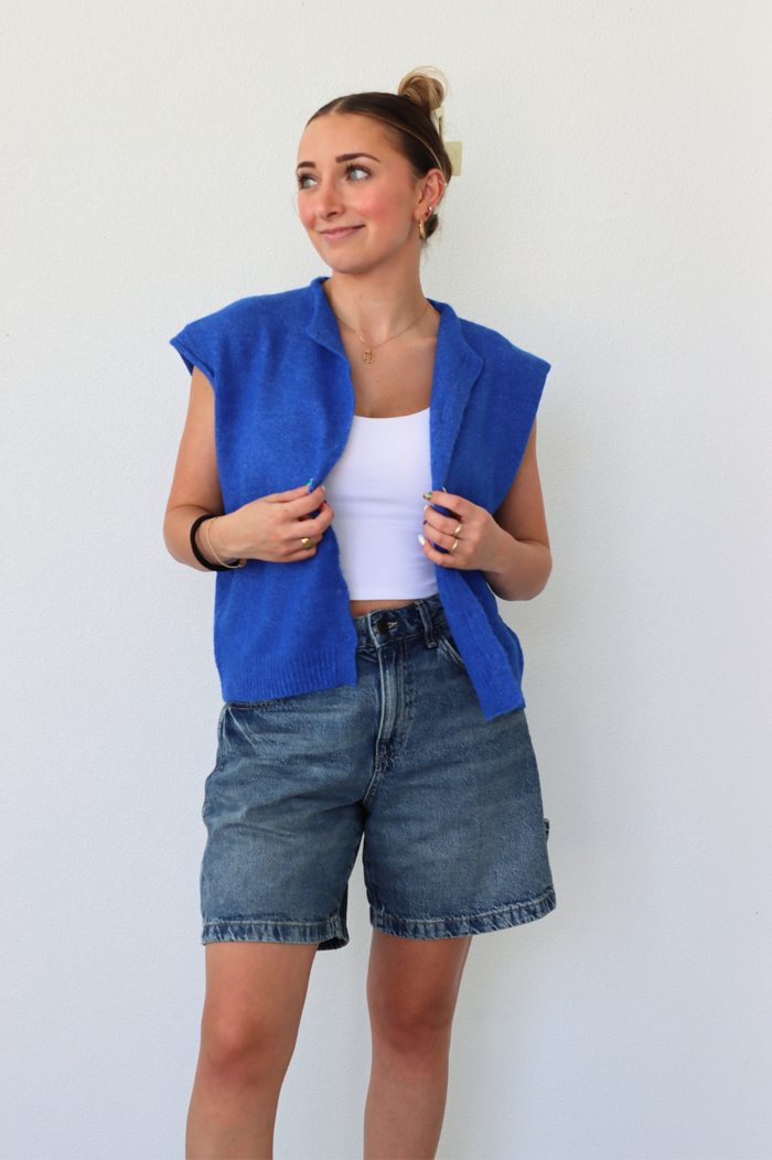 Woman wearing a blue sleeveless top and denim shorts against a white background