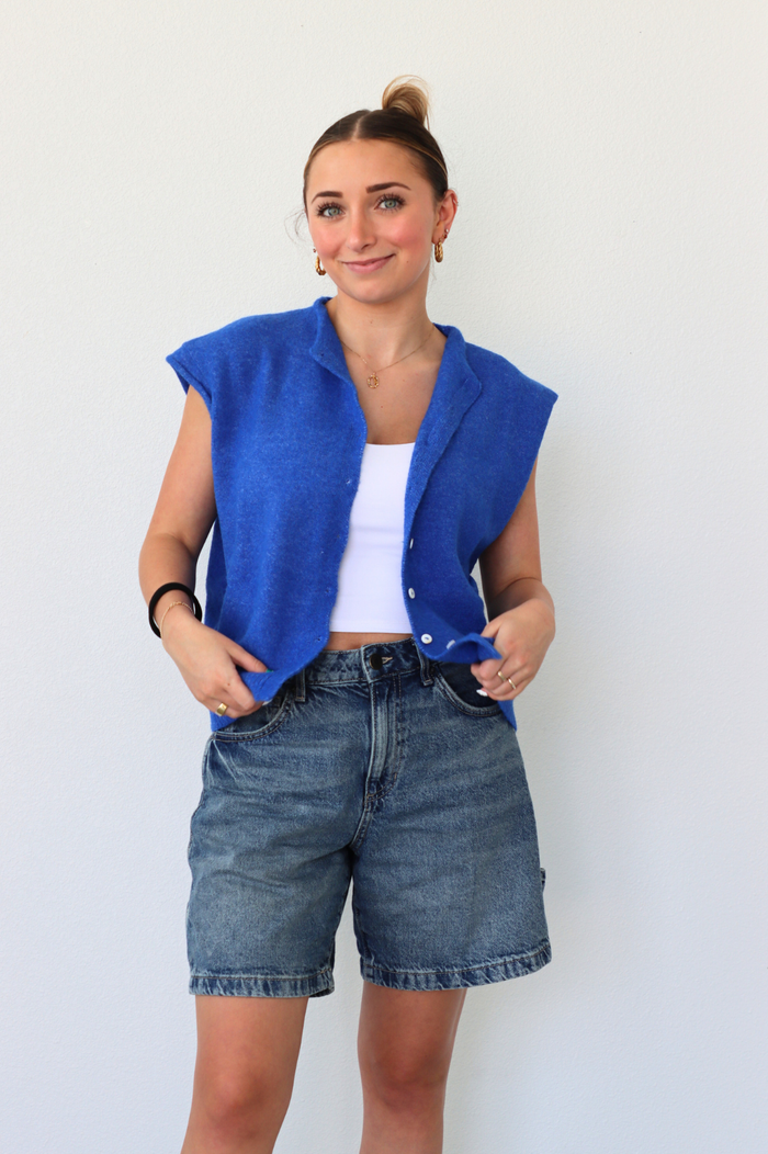 Woman wearing a blue sleeveless jacket over a white top and denim shorts against a plain background