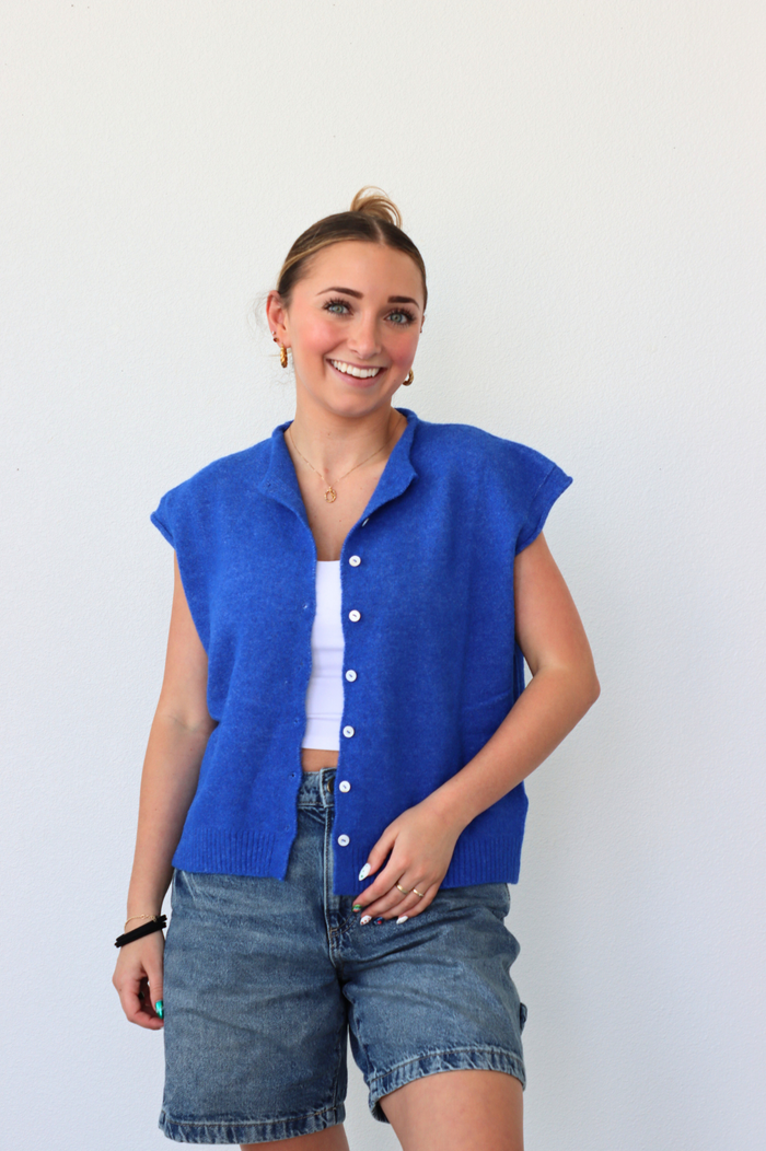 Woman wearing a blue sleeveless vest over a white shirt and denim shorts on a plain background