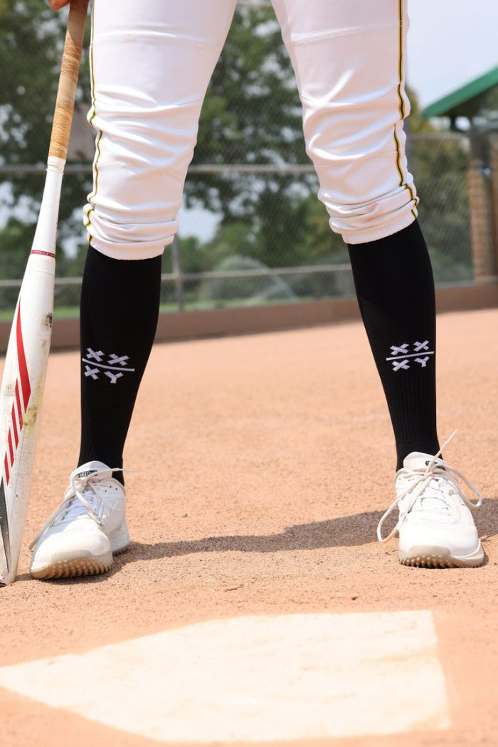 Person on a baseball field wearing black socks with white designs, white shoes, and holding a baseball bat. || Black/White