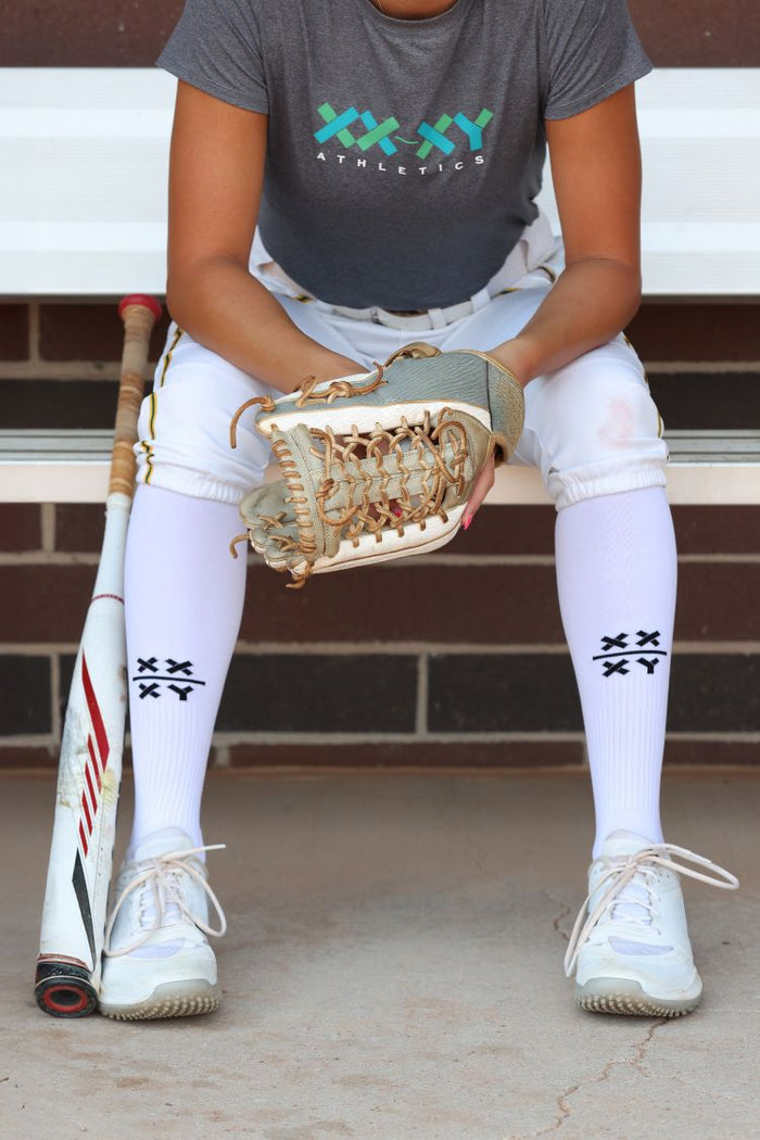 Person sitting on a bench wearing a gray 'XXXY Athletics' t-shirt, white pants, and holding a baseball glove and bat. || White